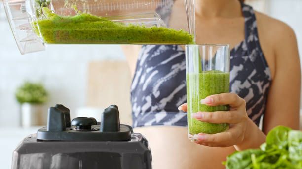 femme sportif faisant le smoothie frais vert sain par le mélangeur pour la boisson saine, y compris le légume et les fruits - green smoothie smoothie nutritional supplement leaf vegetable photos et images de collection