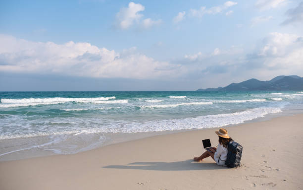 istock A female traveler using and working on laptop computer while sitting on the beach 1303255456