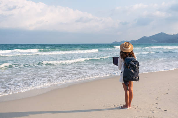 eine reisende, die am laptop unterwegs ist und arbeitet, während sie am strand spazieren geht - nomade stock-fotos und bilder