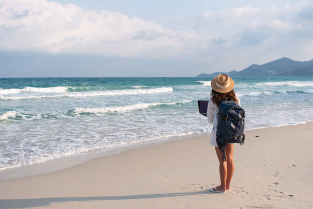 istock a female traveler using and working on laptop computer while walking on the beach 1303255471