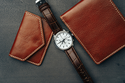 Male watch and leather wallet on dark grey surface close up