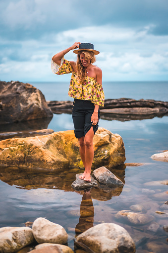 Caucasian blonde girl in a floral shirt, black shorts and straw hat in a natural landscape by the sea and rocks at sunset, lifestyle. Tucked into the water of a natural lake