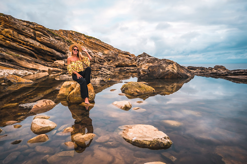 Caucasian blonde girl in a floral shirt, black shorts and straw hat in a natural landscape by the sea and rocks at sunset, lifestyle. Sitting on a rock in a natural pool