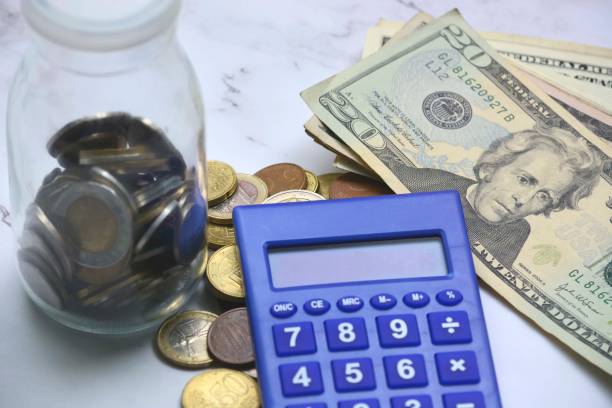 istock Glass jar with multicurrency coins, US dollar bills and calculator white table. Saving and travelling concept 1303285739