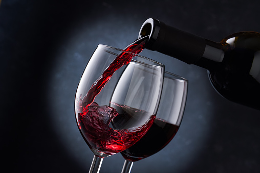 Red wine is poured into a glass from a bottle on a blurry blue background, a stream of red wine from the bottle swirls in the glass, close-up. Free space for text.