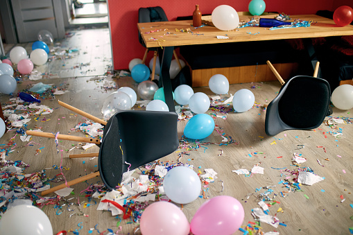 After party chaos, messy in livving room at home, table with pizza and champagne glasses covered with confetti and ballons, chair on the floor at morning after party celebration.