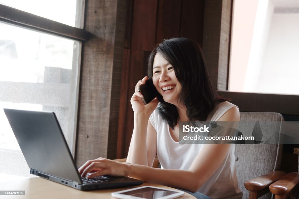 Smiling young Asian woman is calling with smartphone and online working with laptop in living room. Work from home for Covod-19 outbreak concept. Adult Stock Photo