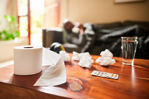 Shot of a medicine with glass of water and tissue paper on table with a sick senior woman lying on sofa in background at home