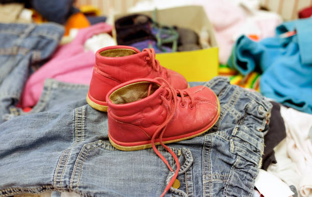 貧困の概念:他の中古服の間のリサイクルショップで子供たちに赤い靴を使用 - 選択的な焦点 - 乳児用衣類 ストックフォトと画像