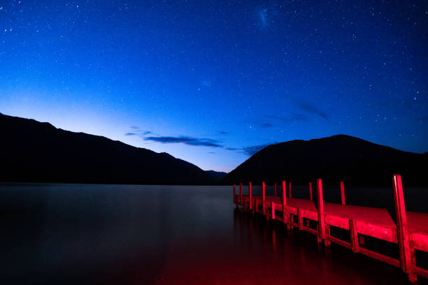 Stars Above Lake Rotoiti, New Zealand In this January 2021 long-exposure photo, stars fill the sky above Lake Rotoiti before dawn. The lake is viewed from its northern shore in Saint Arnaud, New Zealand. The lake is located within Nelson Lakes National Park. This image is a single frame not a composite. The red light on the pier is generated by a headlamp. dusk long exposure nelson rotoiti south island stock pictures, royalty-free photos & images