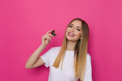 Cute smiling woman in whte shirt is holding chocolate bitten cookie. Waist up studio shot on pink background.