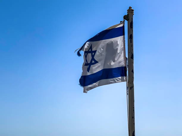 istock Partially torn israeli flag is waving in the wind on blue sky background 1303296877