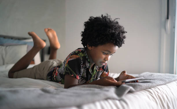 menino deitado na cama jogando jogos em tablet digital em casa - video game child playing leisure games - fotografias e filmes do acervo