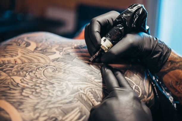 The skin with the style Shot of a tattoo artist hands holding a machine while creating a black and white tattoo on a men back tattooing stock pictures, royalty-free photos & images