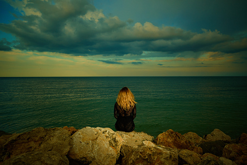 Rear view of blond woman wearing black dress looking at view at the sea with dramatic sky