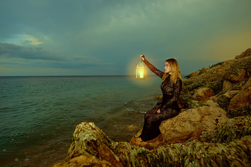 Profile view of woman wearing black lace dress at the sea holding lantern looking at it feeling mystical