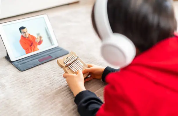 A boy in headphones is playing kalimba with a teacher remotely over a video conference system.  Online training, online classes. Distance education. A fun online lesson on the internet. Modern lifestyle in lockdown at home.