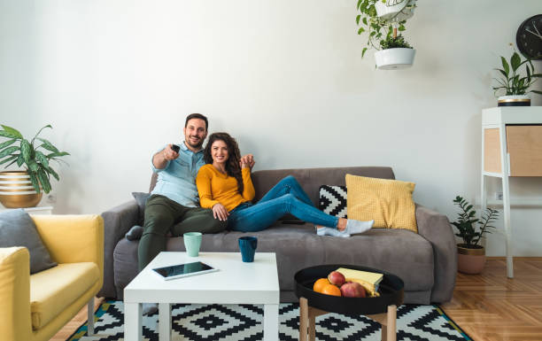 Smiling couple sitting together at home and watching tv Embraced couple watching television while relaxing together on their living room sofa at home part of a series stock pictures, royalty-free photos & images