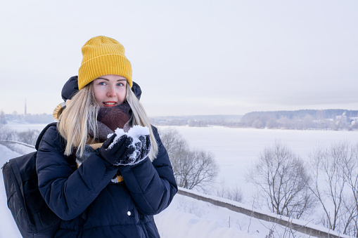 Young smiling woman in yellow beanie and black padded jacket with black backpack is holding snow in her hands against the winter landscape with icy river