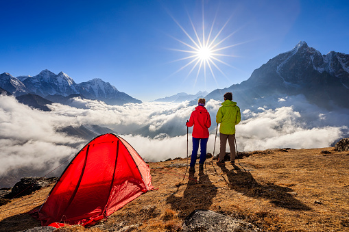 Young woman, wearing red jacket and young man wearing green jacket, are standing on the top of a mountain, next to their tent, and watching sunset over Himalayas .Mount Everest National Park. This is the highest national park in the world, with the entire park located above 3,000 m ( 9,700 ft). This park includes three peaks higher than 8,000 m, including Mt Everest. Therefore, most of the park area is very rugged and steep, with its terrain cut by deep rivers and glaciers.