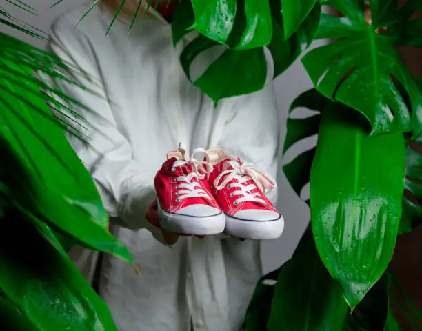 Photo of Woman in white shirt show red gumshoes near palm leaves.