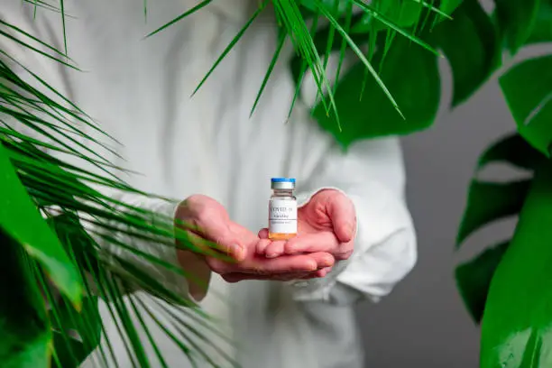 Photo of Woman in white shirt show vaccine in a bottle near palm leaves.