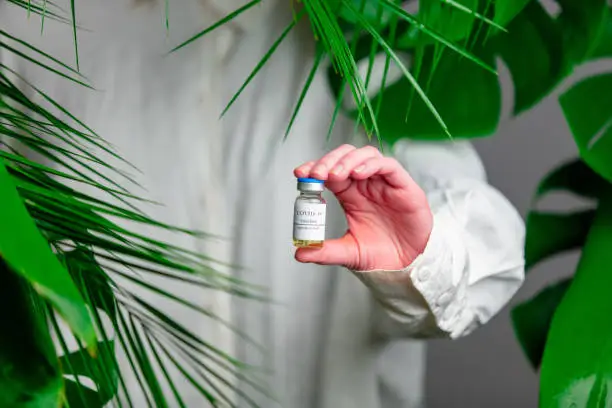 Photo of Woman in white shirt show vaccine in a bottle near palm leaves.