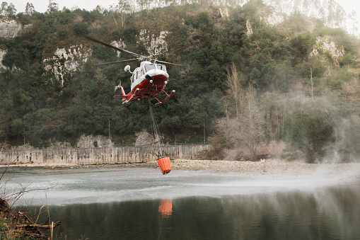 Fire fighting helicopter loading water into a bucket in a river to extinguish a fire nearby