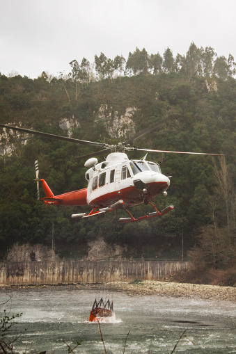 Fire fighting helicopter loading water into a bucket in a river to extinguish a fire nearby