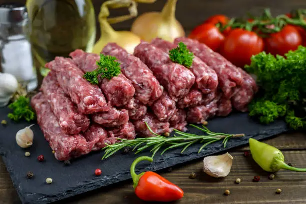 Raw minced meat cevapi ready for barbeque with various fresh vegetables on a wooden table. Selective focus.