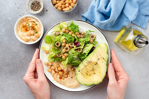 Female hands holding salad buddha bowl with avocado, chickpea hummus and greens. Top view. Clean eating, dieting concept