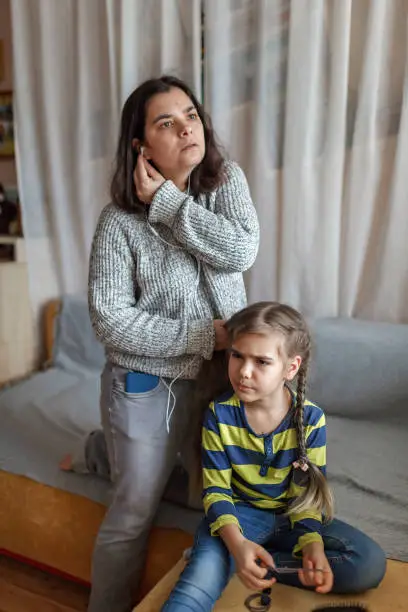 Photo of Mother getting ready daughter for school, combing her hair and listening audio chat at once