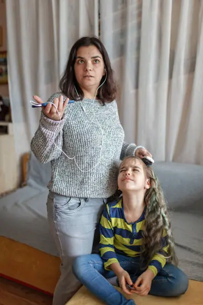 Photo of Mother getting ready daughter for school, combing her hair and listening audio chat at once