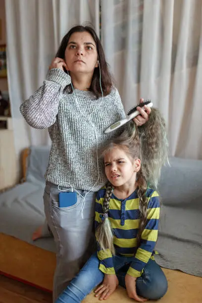 Photo of Mother getting ready daughter for school, combing her hair and listening audio chat at once