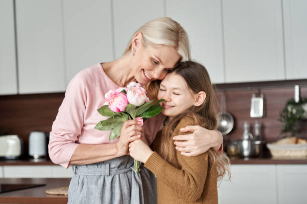 istock Happy beautiful affectionate middle aged mom receiving spring flowers bouquet hugging cute teen child kid daughter standing at home in kitchen enjoying surprise moment celebrating Mothers Day holiday. 1303367741