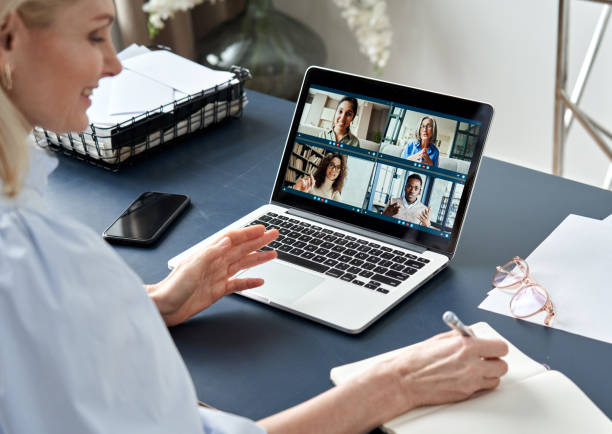 istock Business woman having virtual team meeting on video conference call using laptop computer. Social distance work from home office with diverse people group in remote online chat. Over shoulder view 1303367754