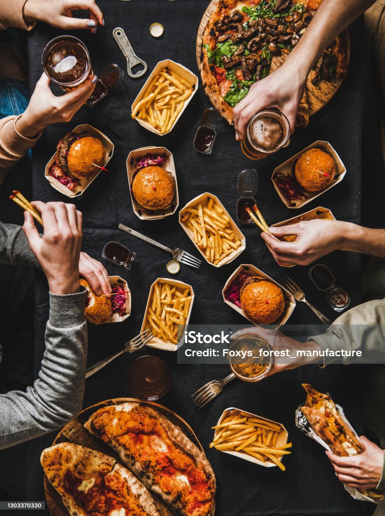 Flat-lay of friends having quarantine home party with fast food Lockdown family fast food dinner from delivery service. Flat-lay of friends having quarantine home party with burger, fries, sandwiches, pizza, beer over dark table background, top view Food Stock Photo