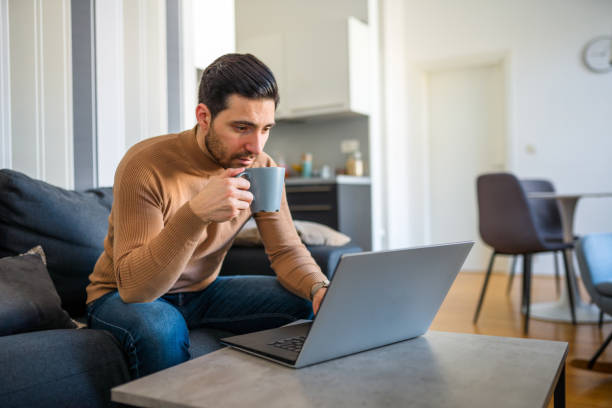 istock Caucasian Man Using Laptop and Drinking Coffee on Sofa at Home 1303372533