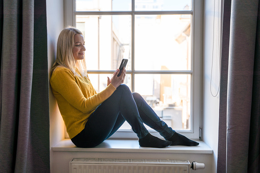 Caucasian mid adult woman sitting on a window sill at home and using mobile phone. She is wearing casual clothes and having online meeting.