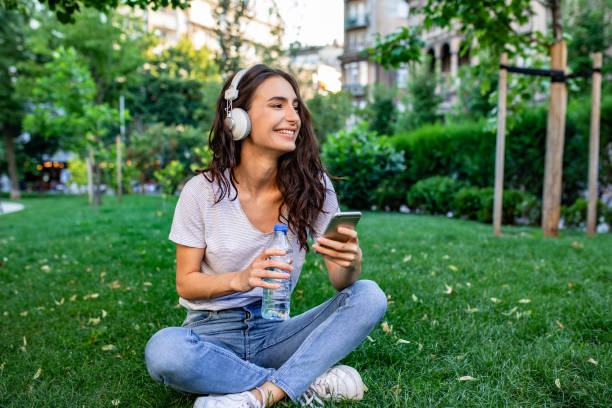 Young woman sitting on the grass in park and relaxing Photo of young woman relaxing, drinking water and using phone green environment friendly stock pictures, royalty-free photos & images