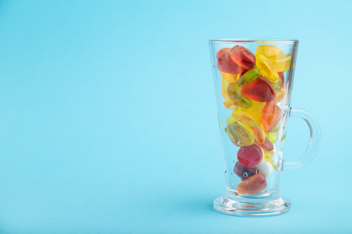 Various fruit jelly candies in drinking glass on blue pastel background. side view, copy space.