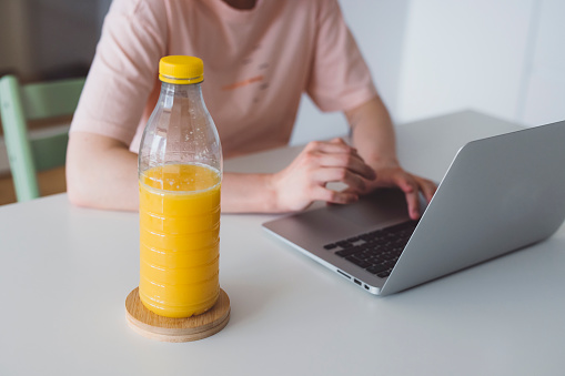Woman working on her laptop sitting by the desk having a plastic bottle full of orange juice next to her. Drinking orange juice while working from home.