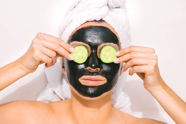 Young beautiful woman with a towel on her head applying black purifying charcoal facial mask and holding cucumber slices on her eyes. Face skin care. Beauty treatments. Spa therapy. Young beautiful woman with a towel on her head applying black purifying charcoal facial mask and holding cucumber slices on her eyes. Face skin care. Beauty treatments. Spa therapy. High quality photo women facial mask mud cucumber stock pictures, royalty-free photos & images