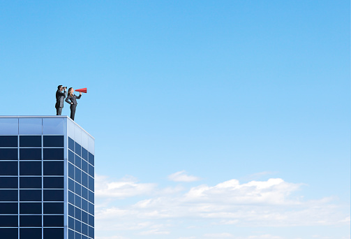 A businessman and a businesswoman stand on top of a tall building. He looks through a pair of binoculars as she shouts through a red megaphone as they try to communicate.