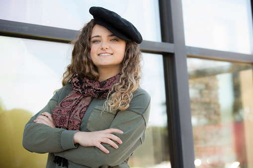 Lovely young woman wearing a stylish Beret stands in front of a shop window.  She looks into the camera with confidence.