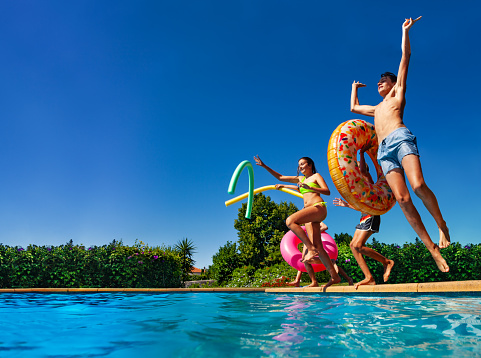Many of happy teenage kids dive in the swimming pool water throw inflatable toys lifting hands have fun