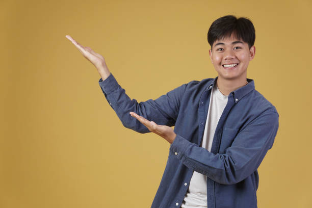 istock portrait of happy handsome young asian man dressed casually presenting with open palms gesture at copyspace. yellow background 1303404377