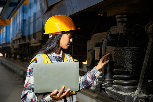 Female engineer wearing protective clothing and helmet is inspecting the availability of the freight train's wheel system. A technician with laptop inspecting work at a railway maintenance workshop.