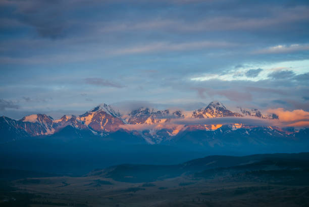 istock Scenic mountain landscape with great snowy mountain range lit by orange dawn sun among low clouds. Awesome alpine scenery with snowy high mountain ridge under blue cloudy sky at sunset or at sunrise. 1303421897