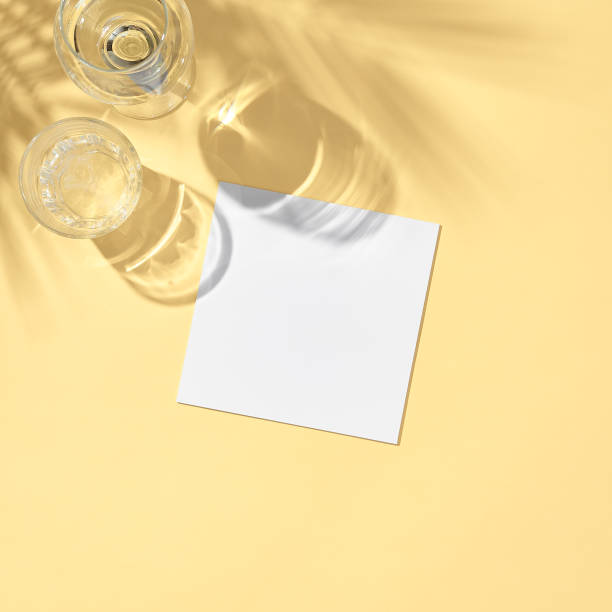 istock Glass with white paper card flat lay. Pastel yellow table in sunlight with palm leaf shadow. Wedding, birthday, tropical summer day concept 1303422954